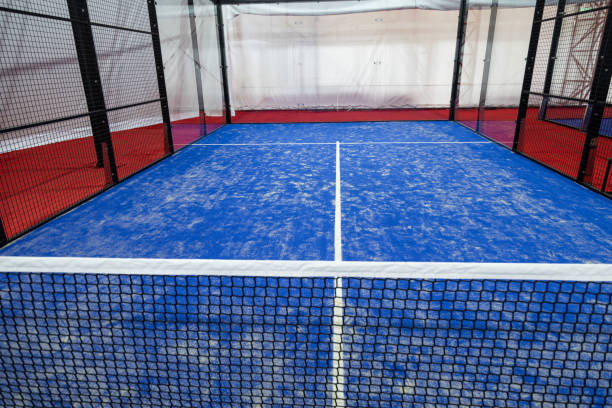 A spacious indoor padel court is designed with a vibrant blue playing surface, white lines marking the center, and netting surrounding the area for safety and play.
