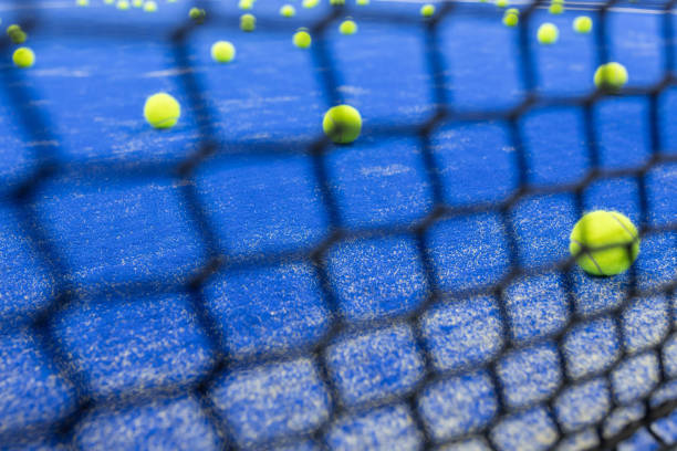 Tennis balls scattered across a blue padel court viewed through a net during practice session in the morning light