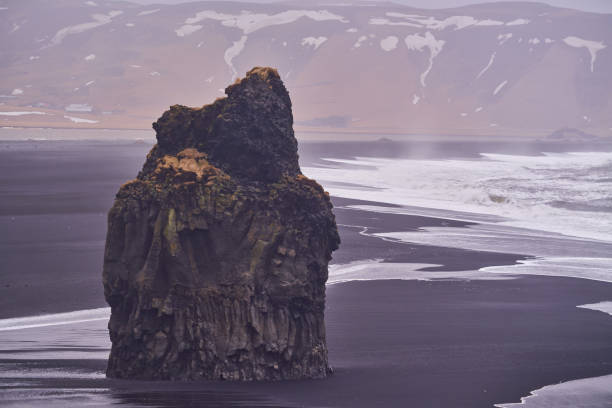 Large rock formation standing on a black sand beach with the rough ocean waves crashing nearby and mountains in the background