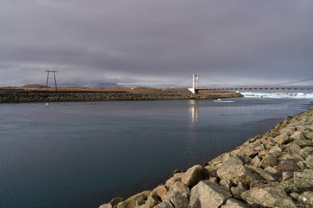 Bridge is crossing a glacier lagoon with mountains in the background on a cloudy day