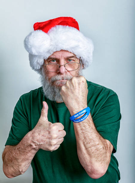 A serious faced Santa Claus hat senior adult man real life, real person, recent cancer and chemotherapy patient is holding a fist up to symbolize strength; giving the thumbs up gesture for success; and wearing two blue colon cancer support wristbands labeled: \