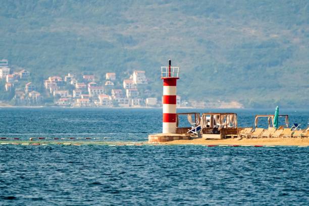 A small red and white lighthouse stands on a sandy pier next to a seaside resort with lounge chairs, overlooking calm waters.