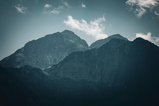 A dramatic view of rugged mountain peaks under a moody sky. The soft light and scattered clouds create a serene yet powerful atmosphere, highlighting the grandeur of the alpine landscape.