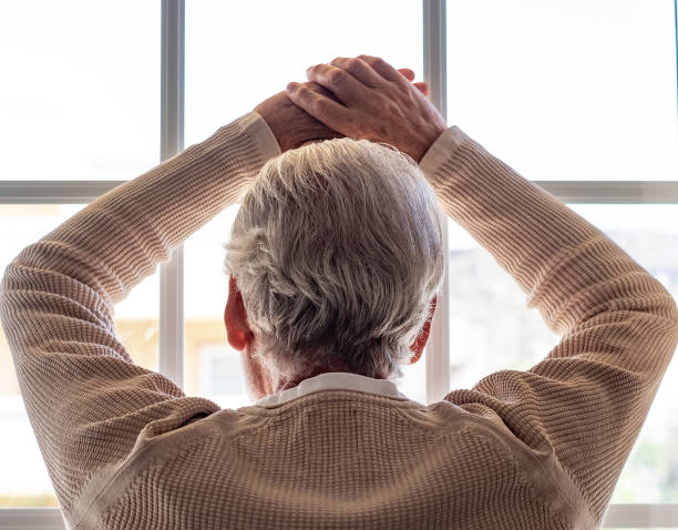 istock Back view of senior gray haired man at home looking out the window, pensive elderly man with hands on glass feeling like a prisoner 2182237607