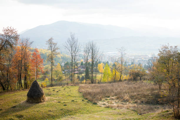 A tranquil autumn scene stretches across rolling hills, with a cozy village nestled in the distance and mountains rising in the background. Golden leaves surround a rustic brown haystack, adding warmth to the landscape.