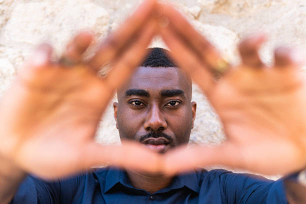 Serious African American adult guy looking at camera while demonstrating triangle shape with hands near fence in street in daylight