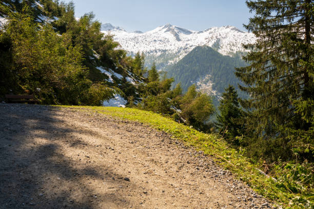 On a sunlit summer morning, a gravel road meanders through the Carnic Alps, while in the backdrop, the Alpine peaks cast blue-hued silhouettes.