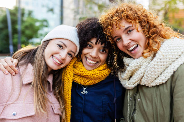 Three young diverse women smiling at camera embracing outdoors in winter stock photo