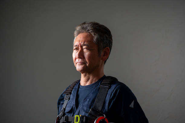 Older Asian man wearing a black t-shirt and cap, standing with arms crossed against a stone wall background, providing space for text or design.