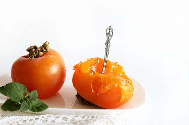 Ripe fresh persimmon fruits served in a white plate with a spoon, decoration with fresh mint leaves on white background. Autumn fruit.