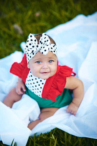 Portrait of a beautiful baby girl in a park, dressed in a beautiful polka dot dress, sitting on a white sheet on the grass.