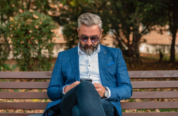 istock A handsome stylish mature business man using mobile phone app texting while sitting on the park bench. 2182353925