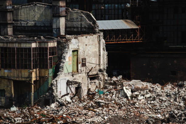 A partially demolished industrial building stands amidst rubble, with rolling hills and other industrial structures in the background. The image reflects the decline of industrial sites and the transformation of landscapes.