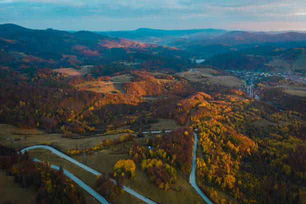 An aerial view of a curved mountain road winding through a vibrant autumn forest. The landscape is illuminated by warm sunlight, highlighting the rich colors of the fall foliage and the scenic route through the hills.