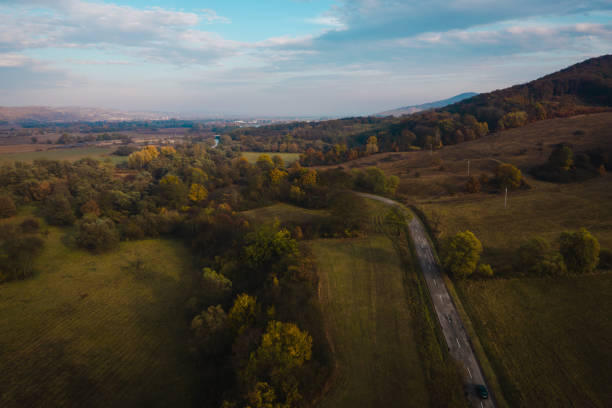A scenic rural landscape with a winding road cutting through open fields, surrounded by trees and gently rolling hills. The late afternoon light enhances the autumn colors, adding warmth and tranquility to the countryside scene.