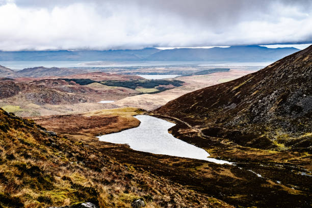istock The landscape features a peaceful lake nestled between hills, reflecting the overcast clouds above, with expansive fields leading to distant mountains under a dramatic sky. 2182455630