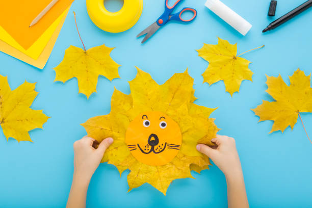 istock Little child hands creating beautiful smiling lion head shape from yellow maple leaves and paper on pastel blue table background. Making autumn decorations. Point of view shot. Closeup. Top down view. 2182460116