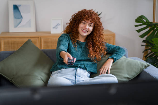 istock Young woman watching tv and holding remote control on sofa 2182466635