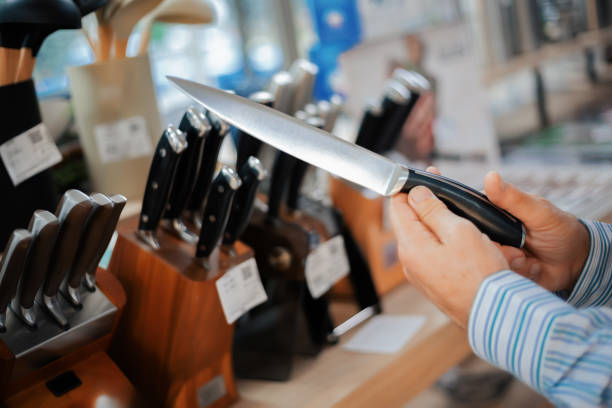 Close-up of hands holding a kitchen knife in a store, examining its blade and design among various knife sets on display.