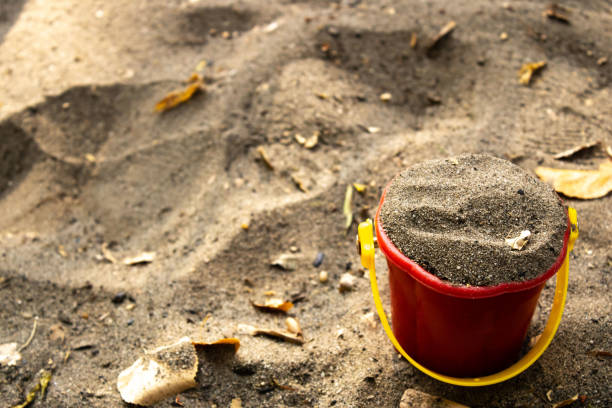A small red bucket filled with sand sits on a sandy playground surface, capturing a simple moment of childhood play. Fallen leaves and scattered debris add to the autumn setting.