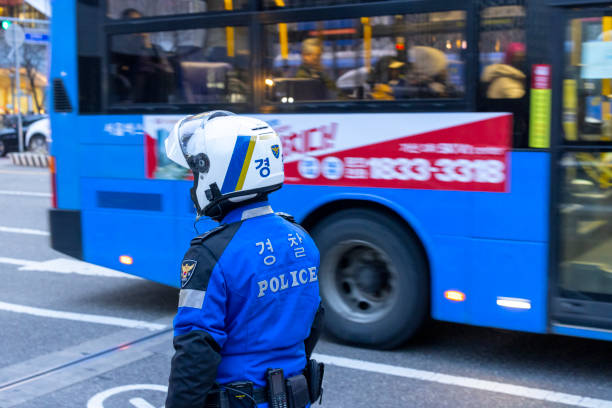 Seoul Police Seoul, South Korea - March 5th 2024: A South Korean police officer in blue uniform and helmet stands watch near a moving city bus on a busy Seoul street, maintaining public safety amidst urban traffic. seoul bus stock pictures, royalty-free photos & images