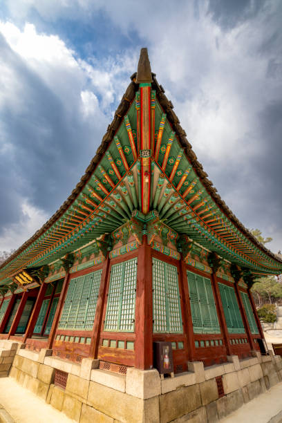 Seoul, South Korea - March 7th 2024: A beautifully detailed traditional Korean building with intricate wooden and painted designs is seen against a dramatic cloudy sky. The architectural style reflects Korea's cultural heritage and artistry.