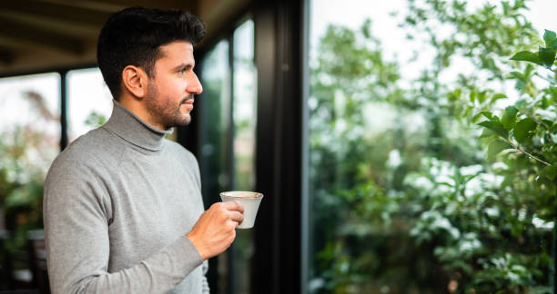 istock Young man drinking coffee and looking out the window at home 2182485855