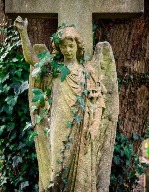 A stone cross in a graveyard, bearing an angel with raised hand. The angel is wearing an ivy head dress. Beckenham Cemetery, Elmers End, London Borough of Bromley.