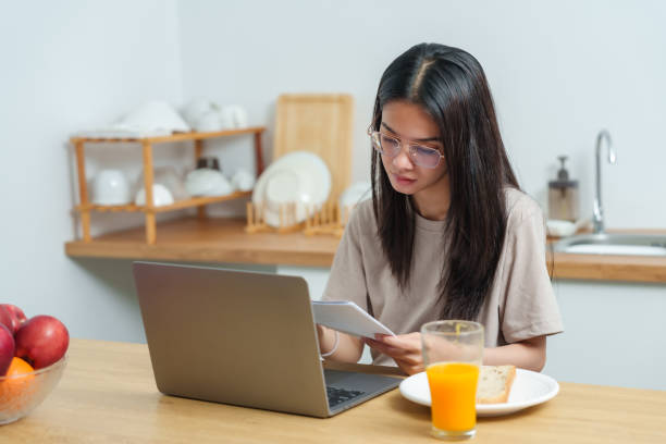 istock Young Asian woman concentrating on document while working on laptop at breakfast table, with orange juice and bread, showing a blend of work focus and calm morning routine. 2182490642