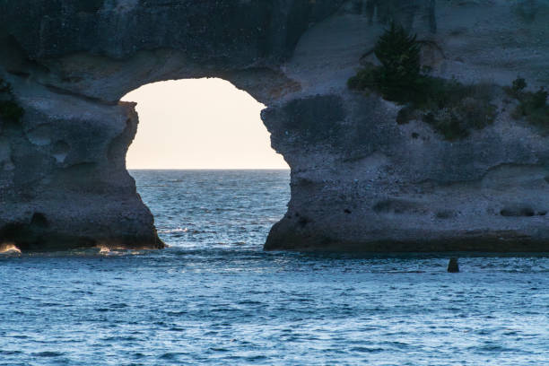 A stunning view of a coastal rock formation with a natural archway framing the tranquil sea beyond. The gentle waves lap against the rock, while the sunlight casts a soft glow on the water, creating a serene and picturesque scene that captures the beauty of nature.