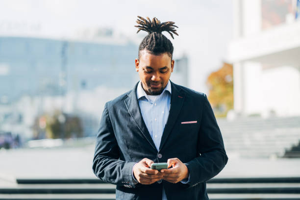 istock Confident, smiling black businessman with dreadlocks in casual blazer standing outdoors on sunny day in urban setting. Business man is using smart phone. 2182516552