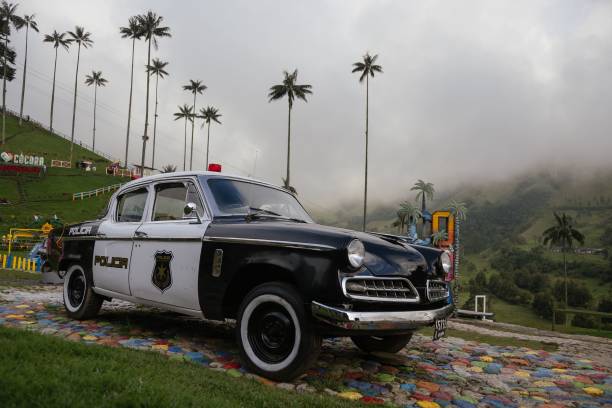 Vintage police car parked on colorful cobblestones with tall palm trees and misty mountains in the background in Cocora Valley, Colombia.
