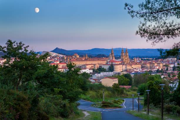 A scenic view of Santiago de Compostela, showcasing the city's skyline with a full moon rising above, framed by lush greenery and distant mountains.