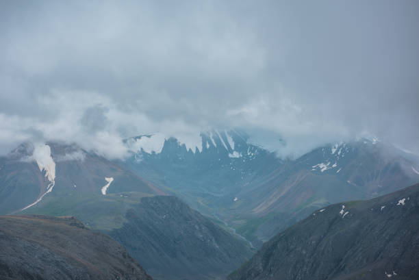 istock Large snow mountain range silhouette in rainy low clouds under gray sky. Mountain stream in green valley in light transparent mist. Hills and rocks in bad weather. Low cloud on big snowy mountains. 2182549315