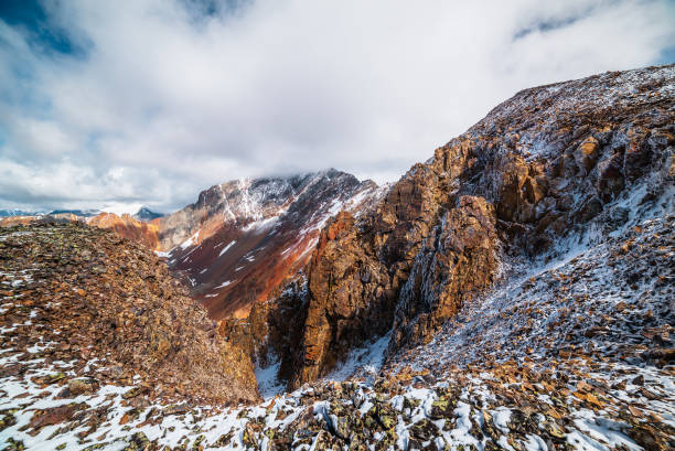 Scenic dizzying top-down view from pass into abyss. Top view to snow-covered stony precipice edge with snowy steep rocky couloir in sunlight. Large mountain range in freshly fallen snow in low clouds.
