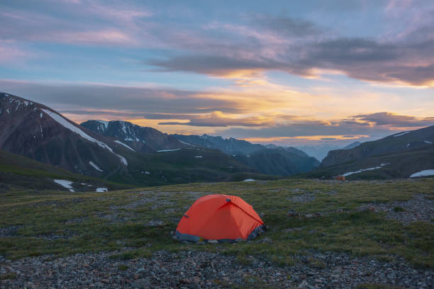 istock Orange tent on grassy stony pass with view to mountain range silhouette far away under clouds in vivid sunset color sky. Scenic alpine landscape with red tent in mountains under colorful cloudy sky. 2182549348