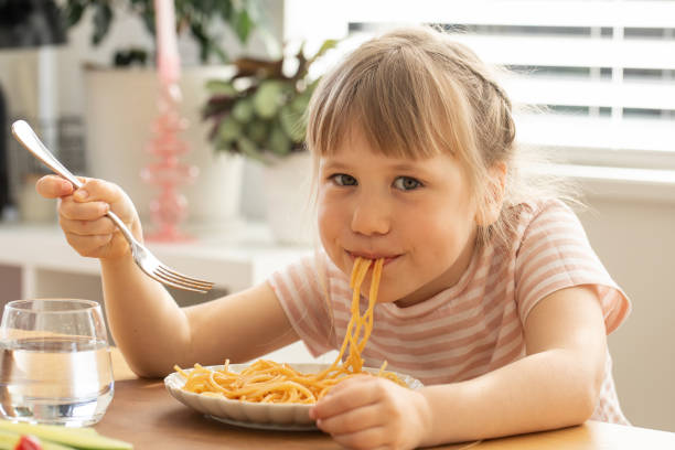 istock Happy little girl eats spaghetti sitting at kitchen table 2182559943