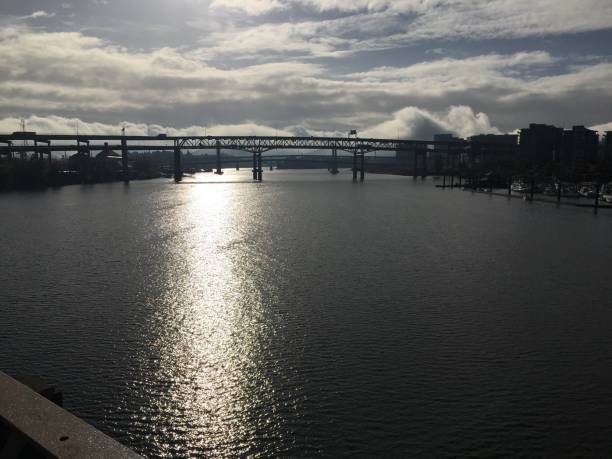 A view of a bridge spanning the Willamette River in Portland, Oregon, with reflective water surfaces under a cloudy sky. The scene includes urban structures and greenery along the shoreline.