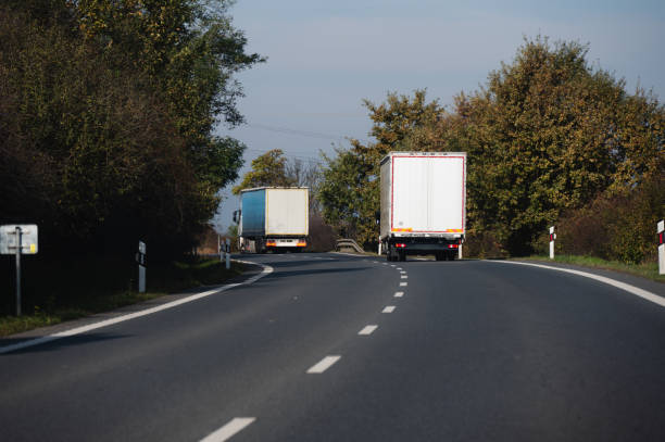 Two large trucks travel on a rural road bordered by trees and bushes under a clear sky. The scene captures a moment of transportation in the countryside, highlighting logistics in a natural setting.