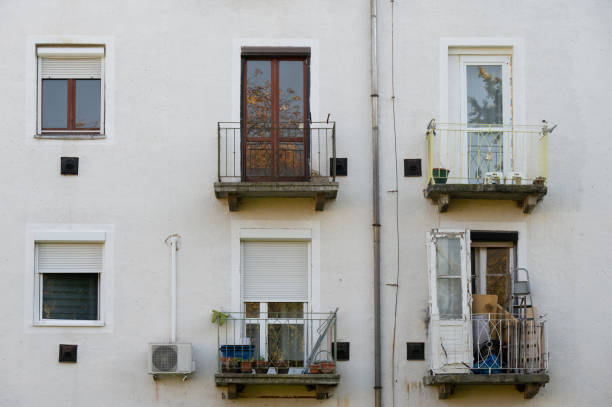 A set of small balconies on an aged apartment building, with various personal items and plants visible. The worn appearance and scattered belongings reflect everyday urban life in a residential setting.