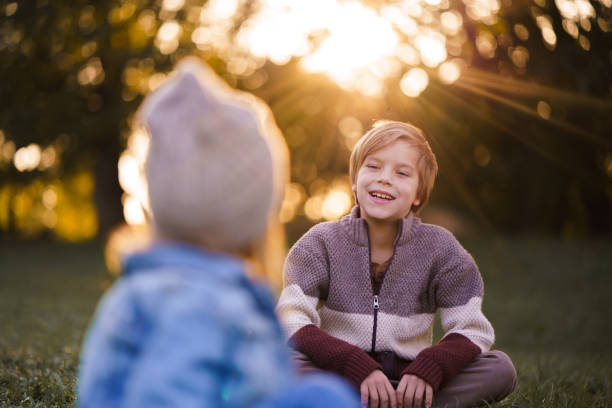 Friendly siblings sit on stone bench resting in tree shadow on summer holidays. Two blond brothers in shorts enjoy spending time together on sunny day