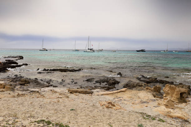 Formentera’s rugged rocks meet crystal-clear waters, with boats gently floating in the background—a stunning scene of natural beauty and Mediterranean charm.