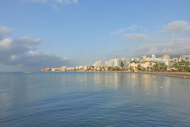 Calm coastal cityscape of Benalmadena Costa, village of Malaga, Andalusia, Spain. Blue water and beachside buildings under a partly cloudy sky, showing a serene Mediterranean shoreline