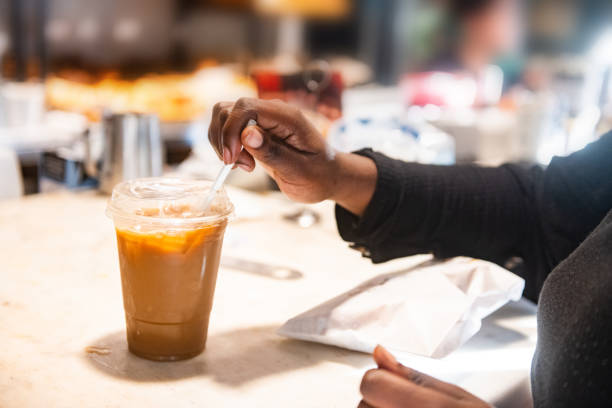 This is a close up photograph of an unrecognizable African American young woman’s hands getting an ice coffee and a snack at the take out counter of an outdoor cafe.
