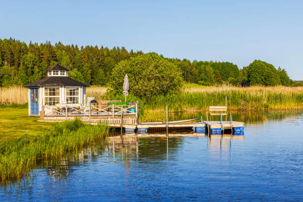 istock Small lakeside gazebo with wooden pier, surrounded by lush greenery under clear blue sky. 2182656410