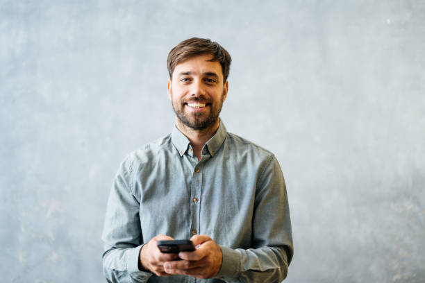 istock Smiling man posing confidently with casual attire against a plain blue background and sending text messages on the smart phone 2182680391
