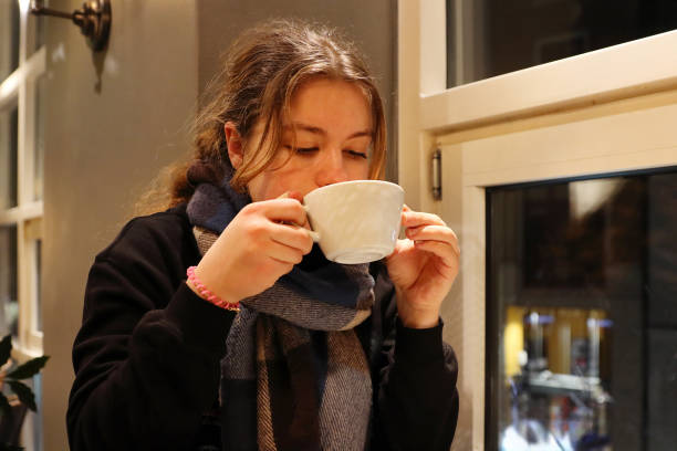 A young beautiful woman sips coffee from the white cup, enjoying a warm drink by a cafe window in the coffee shop.