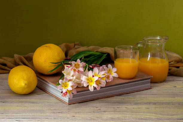 Still life with gentle freesia, lemons and orange juice in a glass and a jug standing on a book on a wooden table close-up