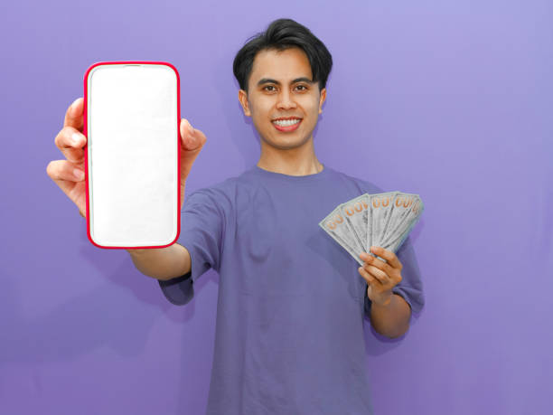 istock A smiling young Asian man displays a blank mobile phone screen while holding banknotes, set against a vibrant purple background. 2182730906