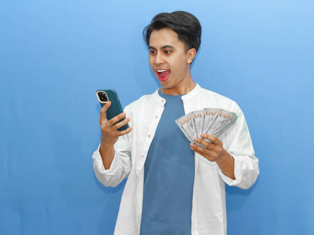 istock A young Asian man looks surprised while using a mobile phone and holding banknotes, set against a vibrant blue background. 2182730947
